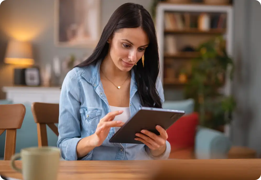 A woman with long dark hair sitting at a table and focused on a digital tablet in a cozy living room.