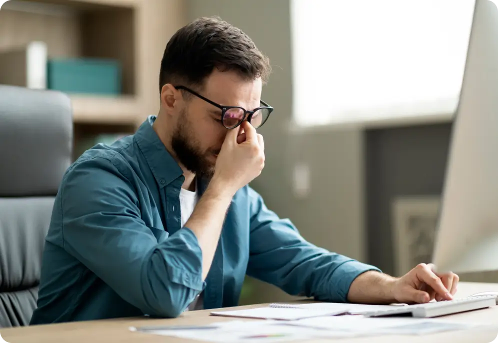 A man wearing glasses sitting at a desk and rubbing the bridge of his nose in fatigue while working on a computer.